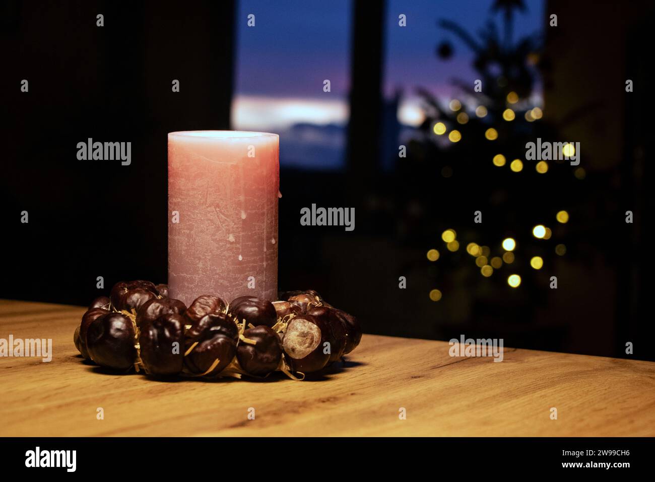 Christmas candle with ring of chestnuts on brown tabel and christmas tree in background near opened window with evening view Stock Photo