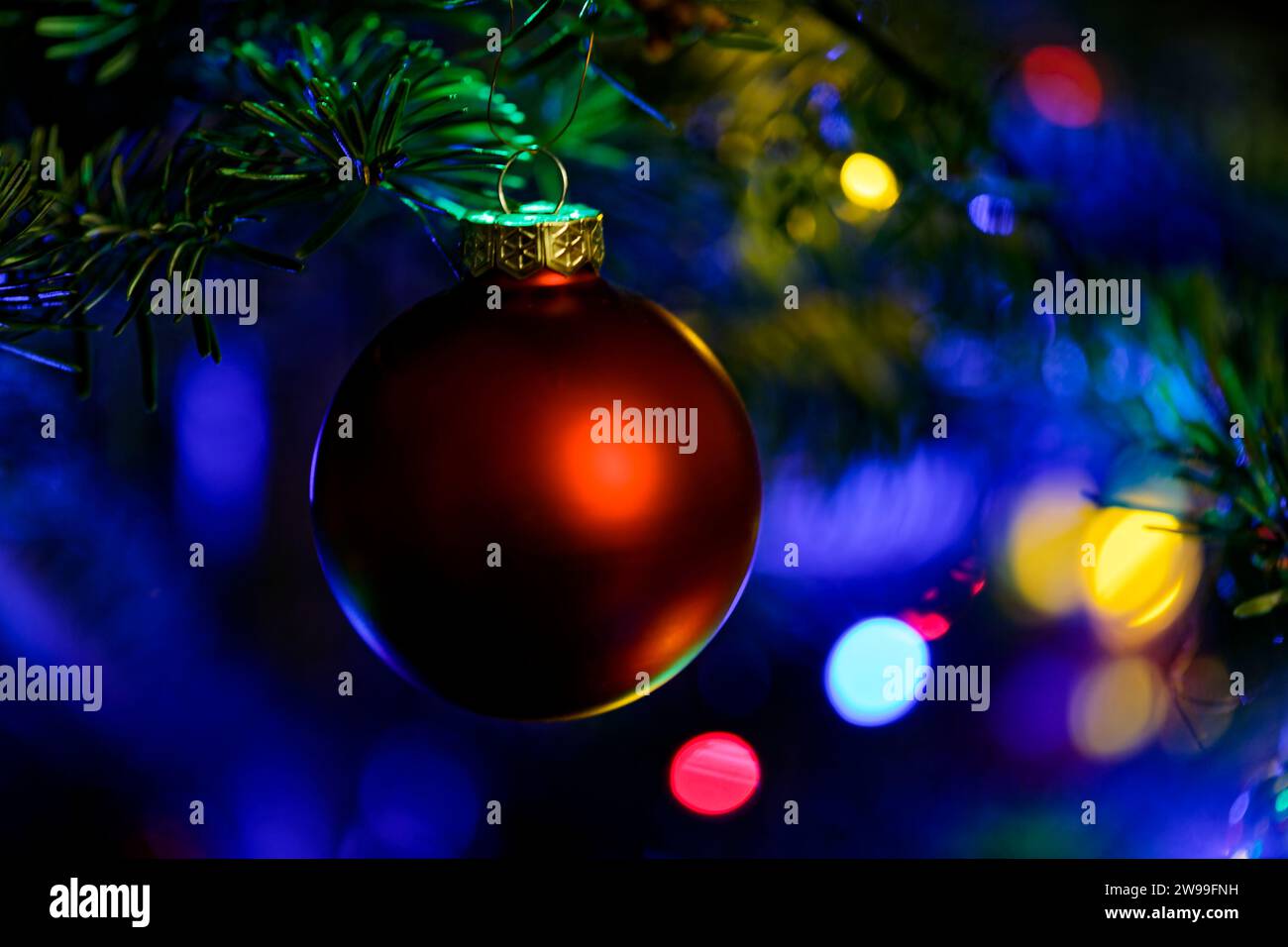 A red Christmas ornament hangs from a tree, illuminated by the vibrant lights of the festive season Stock Photo