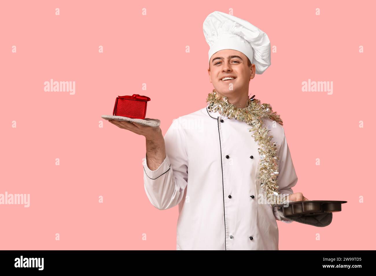 Portrait of young male chef with Christmas tinsel, gift box and baking form on pink background Stock Photo