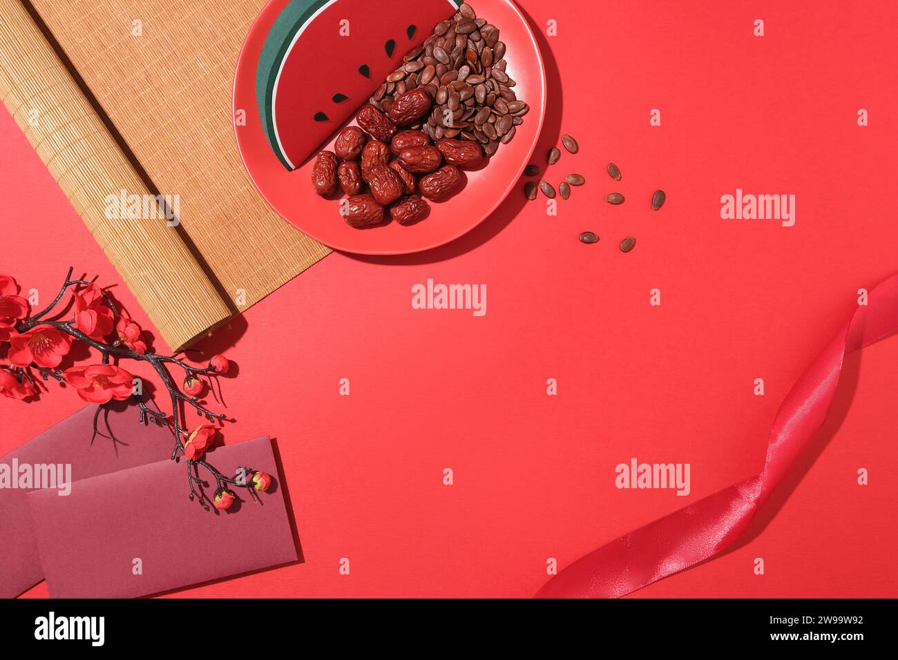 Watermelon, melon seeds and jujube on a ceramic plate, a peach blossom branch, a bamboo curtain and two lucky money envelopes are displayed on a red b Stock Photo