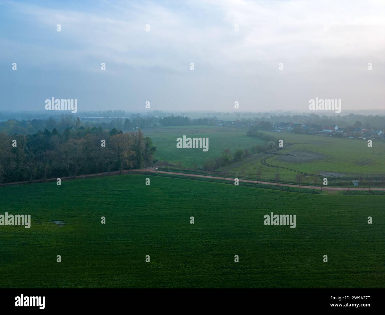 The image captures the serene essence of a misty morning over a sprawling farmland. The soft light of dawn diffuses through the haze, casting a gentle glow over the green fields. The distant horizon is barely visible, shrouded in the morning mist, which gives the landscape an ethereal quality. The curved lines of the terrain and the subtle shades of green create a peaceful, pastoral scene that evokes a sense of tranquility and the slow awakening of the countryside. Misty Dawn Over Gentle Rolling Farmland. High quality photo Stock Photo