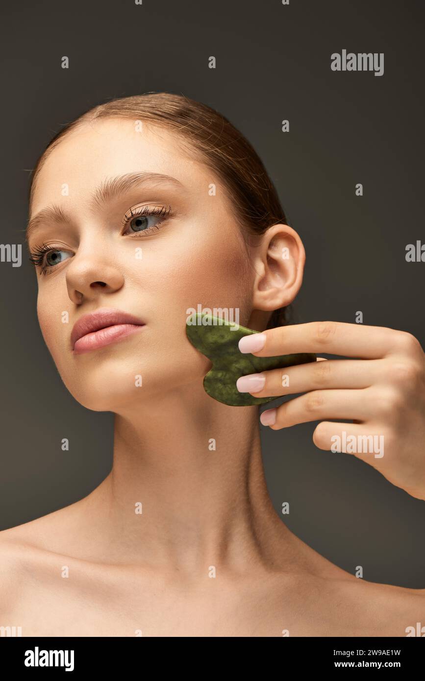 young woman doing face massage with jade gua sha stone on grey
