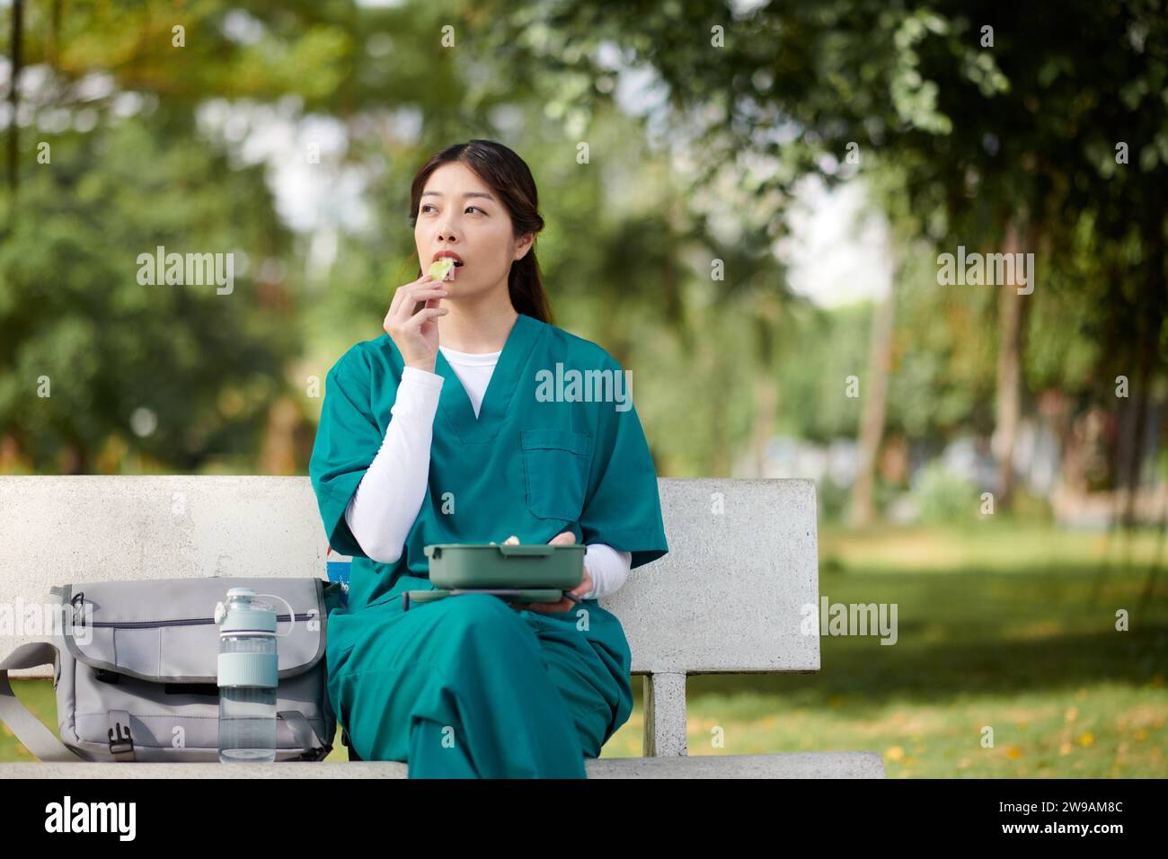 Pensive medical nurse in scrubs sitting on bench in park and eating