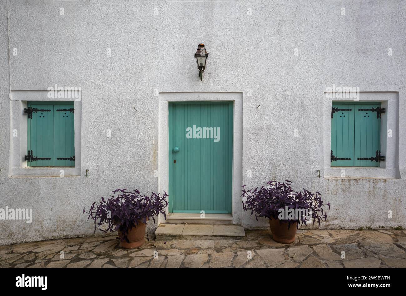 White facade of a house with bright green closed wooden door and window shutters. Lantern above the door. Two pots with violet plants on the stone pat Stock Photo