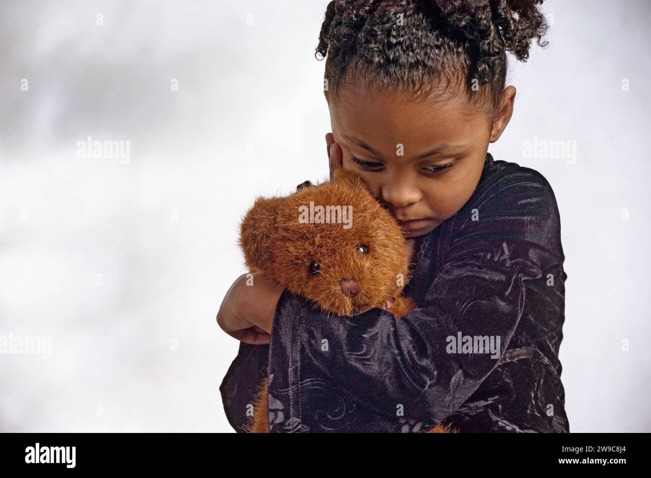 A l very upset ittle African-American girl hugging her teddy bear against a plain background Stock Photo