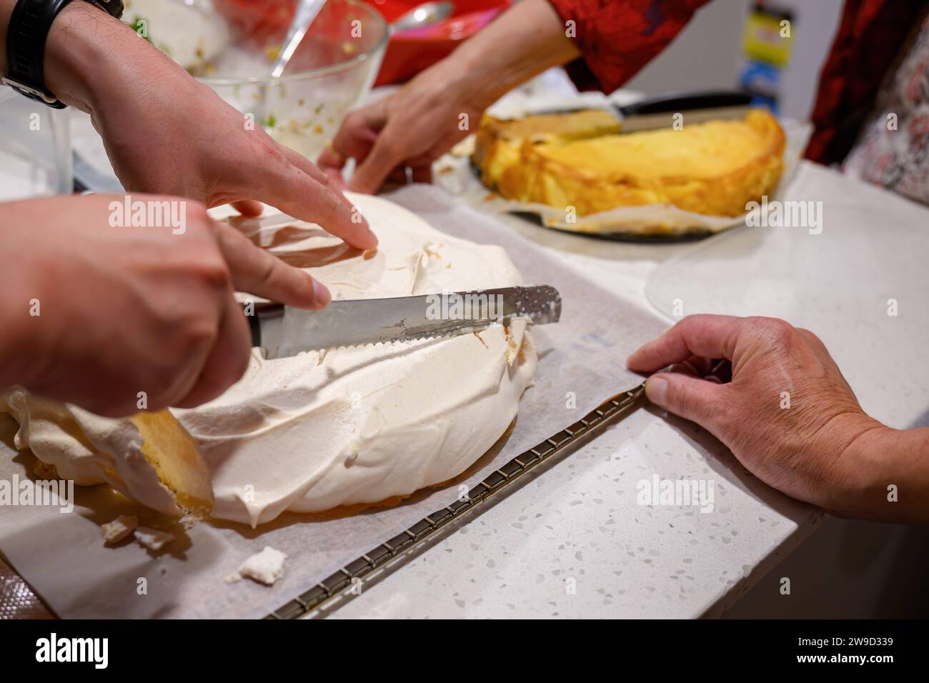 Hands holding knife and cutting an undecorated pavlova on kitchen benchtop. Home cooking and entertainment concept. Stock Photo