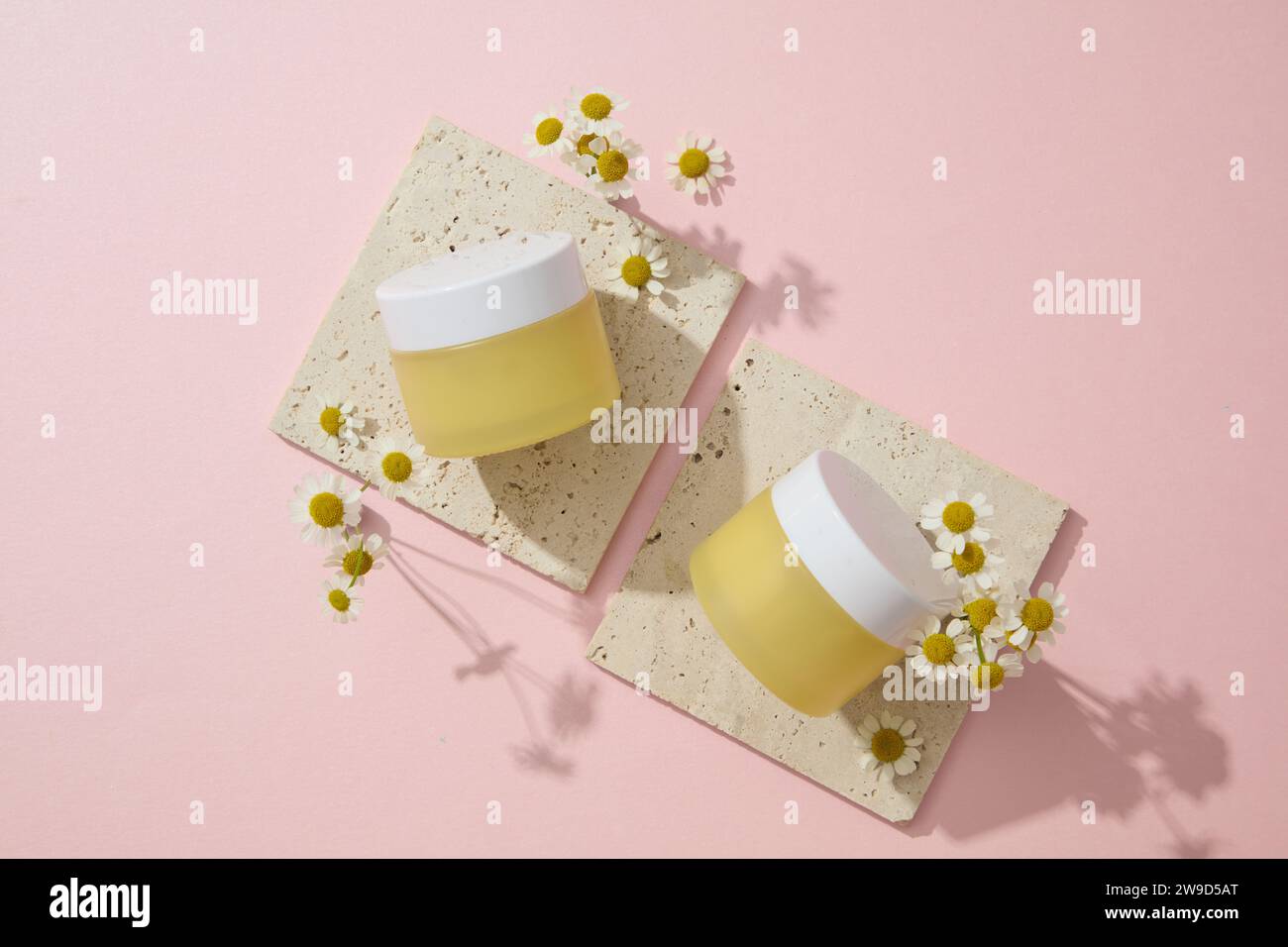 Against the pink background, two yellow cream jars displayed on brick podiums with chamomilla flowers. Mockup scene for advertising cosmetics with moi Stock Photo