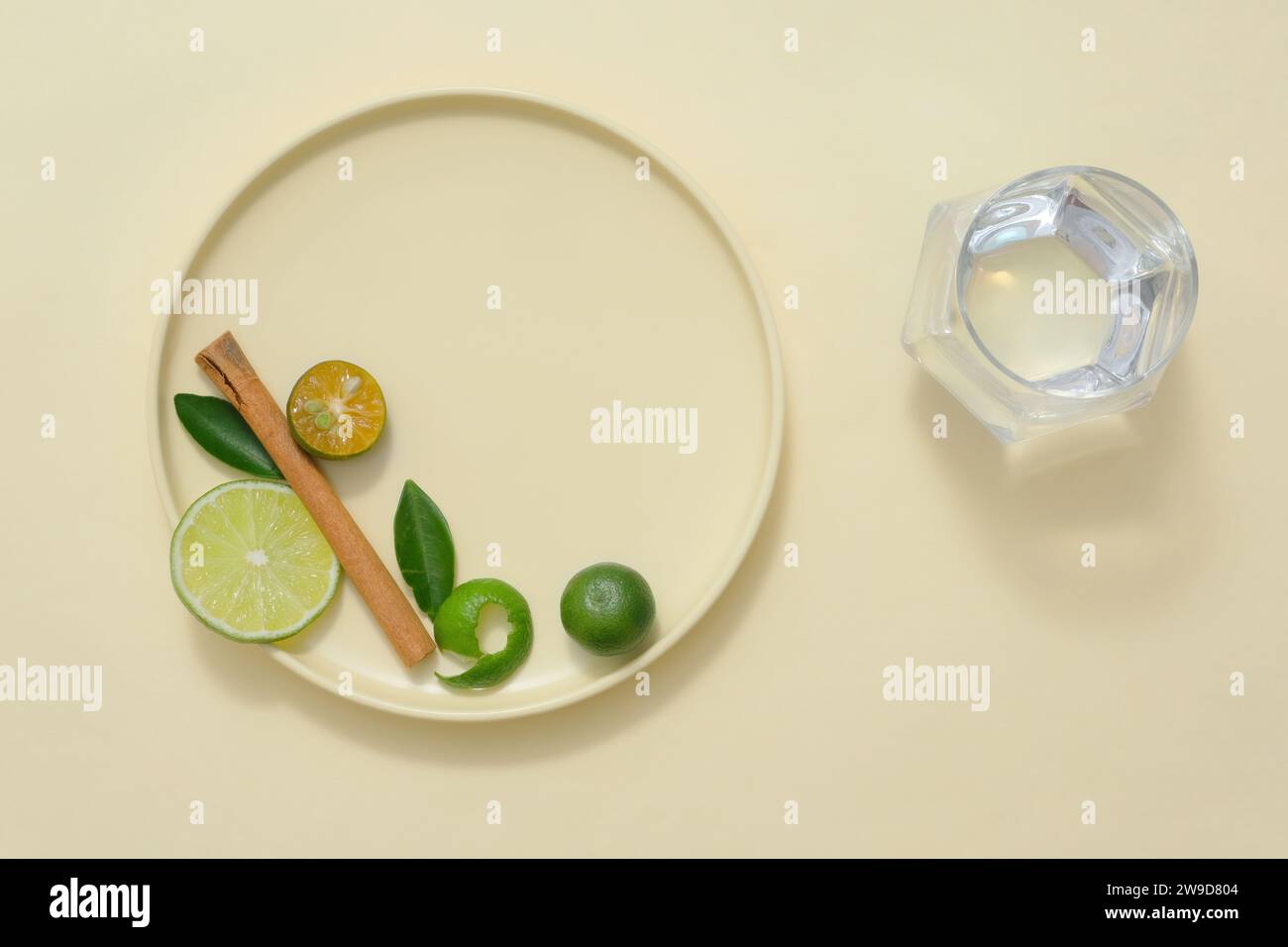 A round ceramic dishes containing slices of lime and kumquat, green leaves and dried cinnamon stick decorated with glass cup of water. Space for desig Stock Photo