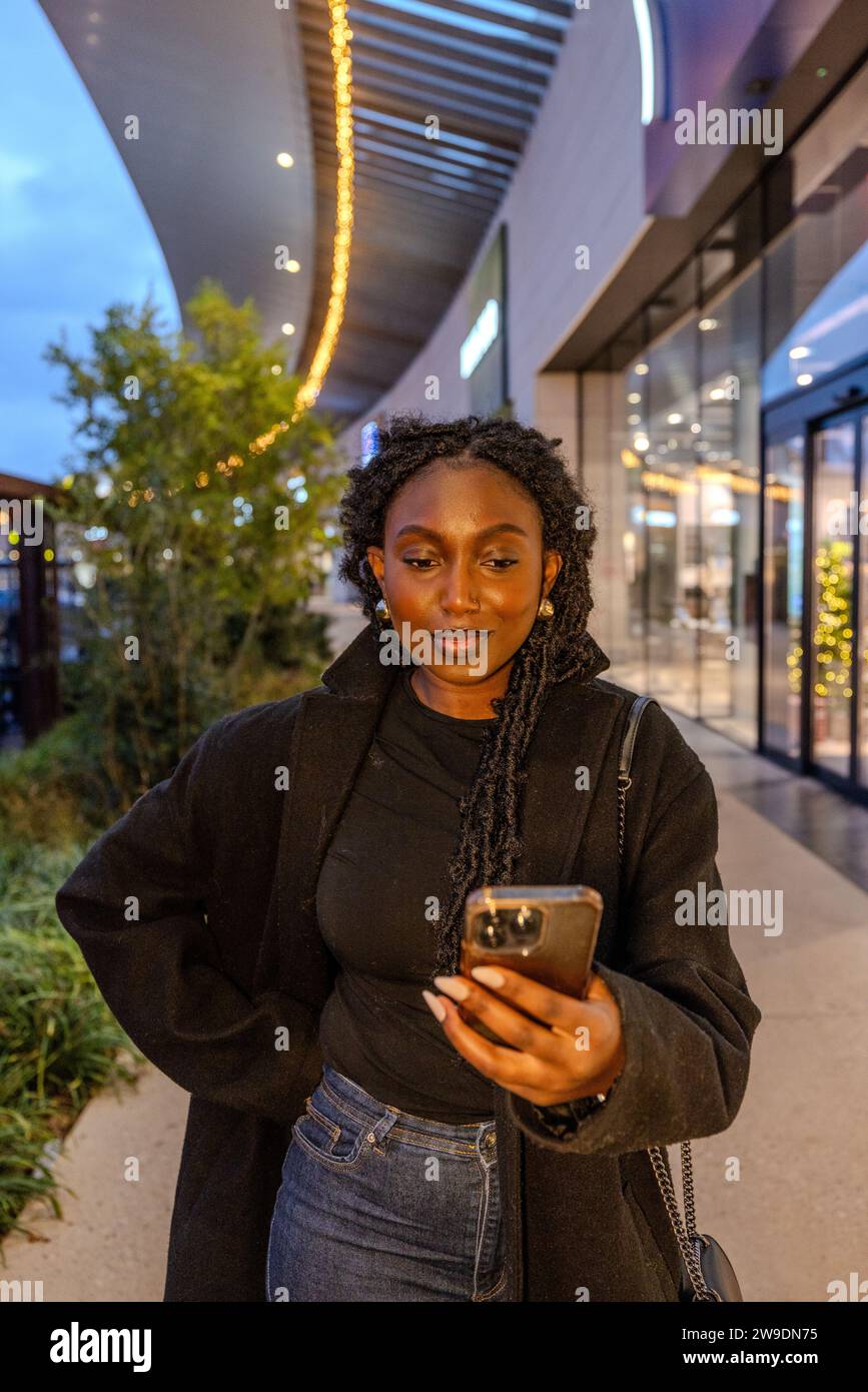 In the soft glow of twilight, a young Black woman is the focal point amidst the architectural lines of an urban shopping promenade. Her attire, a textured black overcoat atop a dark turtleneck, contrasts with the light blue of her high-waisted jeans. She is engaged with her smartphone, perhaps in the midst of digital communication or simply lost in the vastness of the internet. Her stance is relaxed yet assertive, hand on hip, embodying a blend of casual elegance and tech-savviness. This image captures the dichotomy between the traditional physical space and the virtual realms we frequent. Con Stock Photo