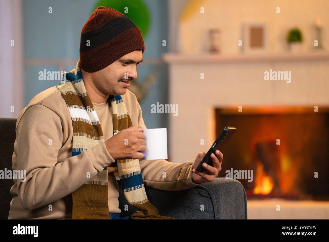 Indian middle aged man in winter wear using mobile phone while drinking tea or coffee at home - concept of cyberspace, social media sharing and Stock Photo