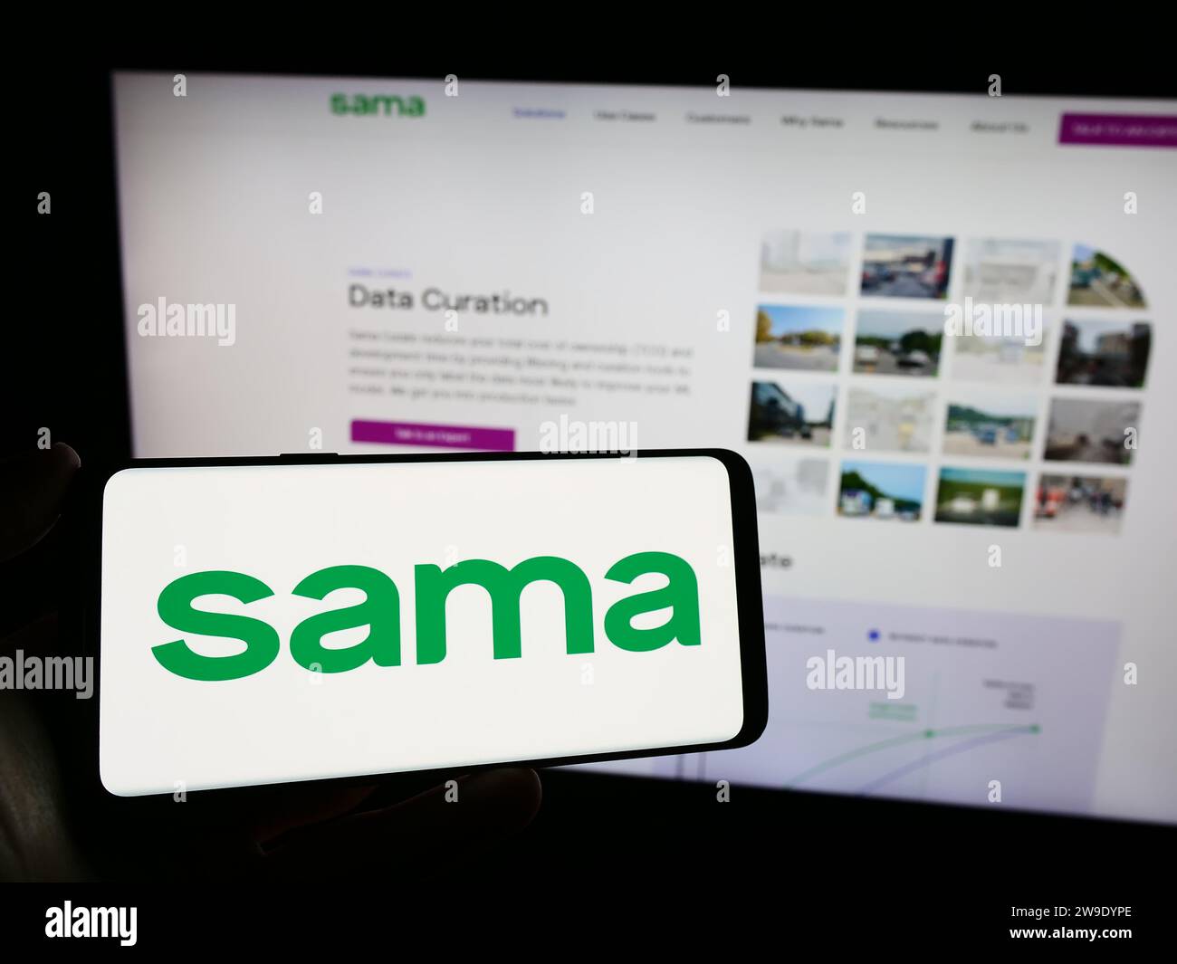 Person holding mobile phone with logo of American company Samasource Impact Sourcing Inc. (Sama) in front of web page. Focus on phone display. Stock Photo