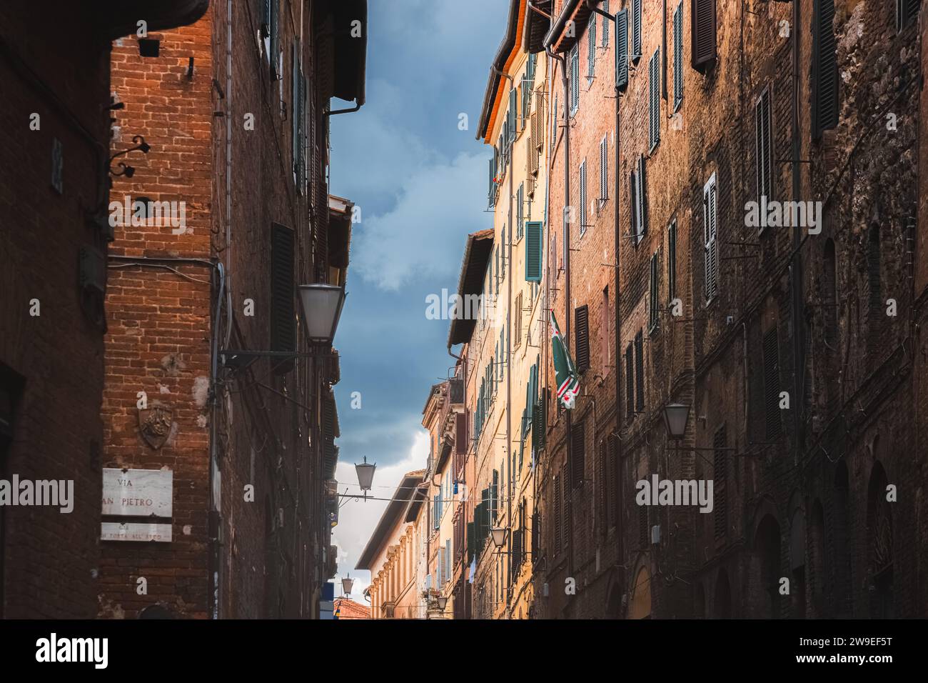 A dark moody sky over colourful residential buildings along a quaint and narrow backstreet in the historic Tuscan old town of Siena, Tuscany, Italy. Stock Photo