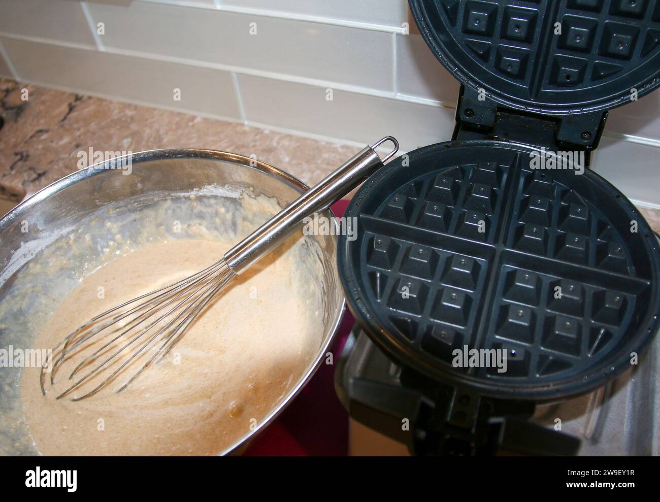 A waffle iron with a whisk inside of a silver bowl filled with waffle mix. Stock Photo