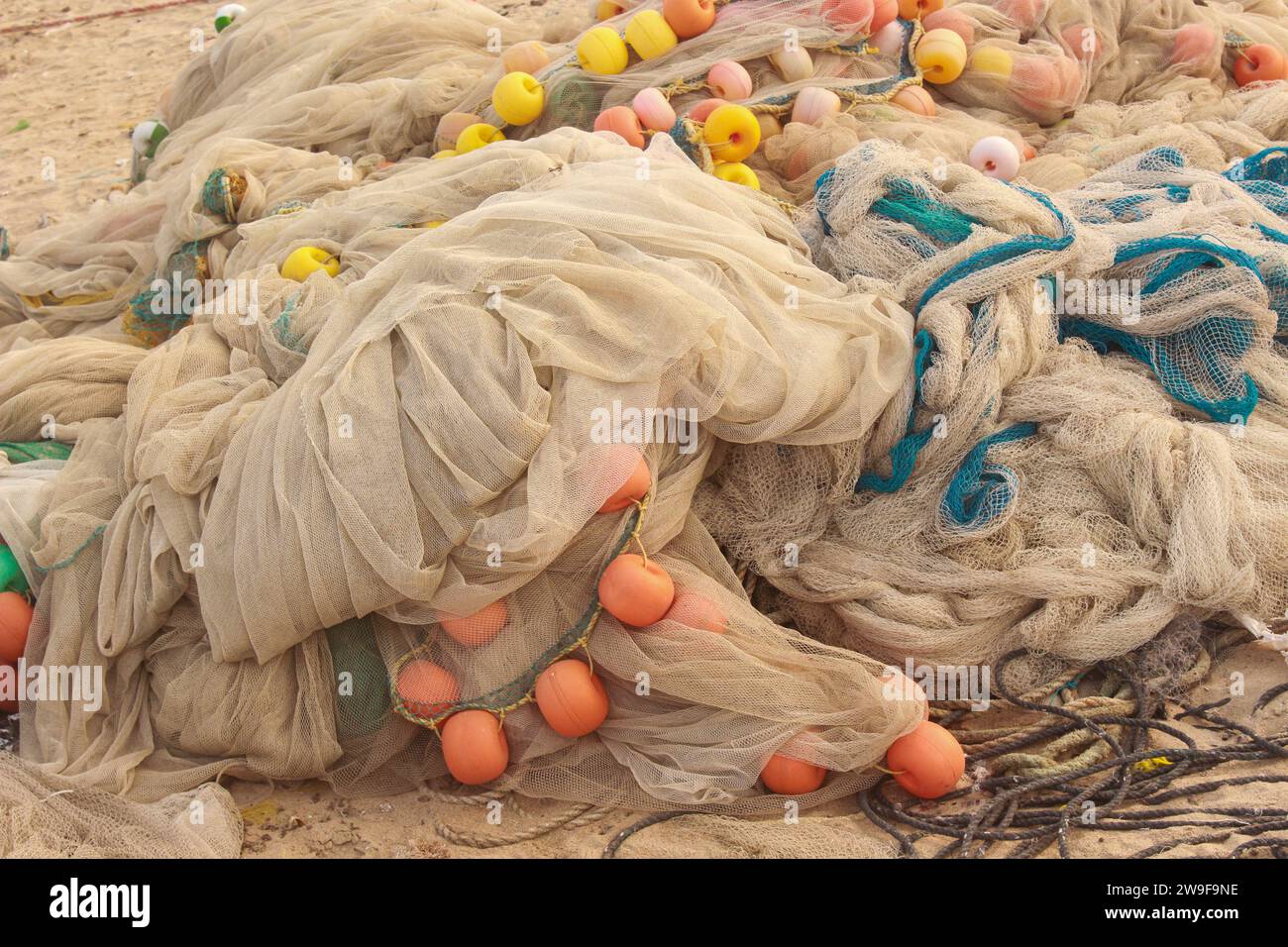 A pile of colorful fishing nets and buoys sits on a sandy beach. The nets are a mix of blues, greens, and oranges, and the buoys are red, yellow, and Stock Photo
