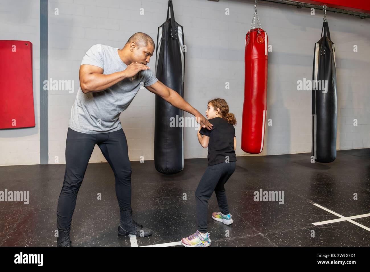 Horizontal photo elementary girl, dressed in tights and black t-shirt, warming up with her black mid adult teacher at a boxing school. Sport, recreati Stock Photo