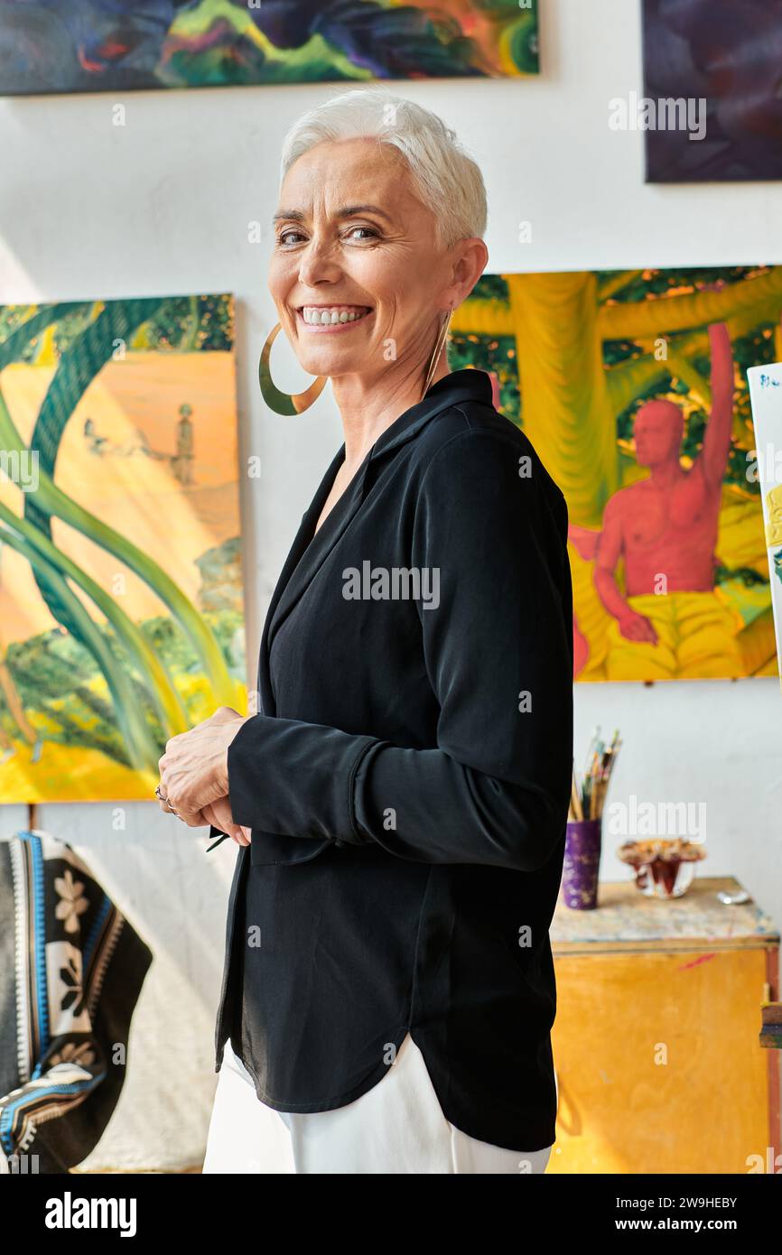 stylish and cheerful middle aged woman artist looking at camera near ...