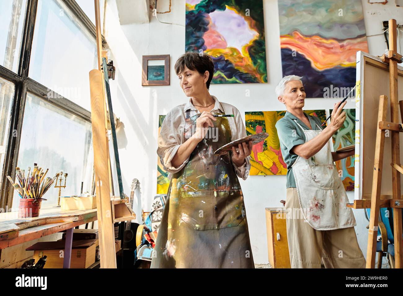 creative mature female artists in aprons pointing on easels in art