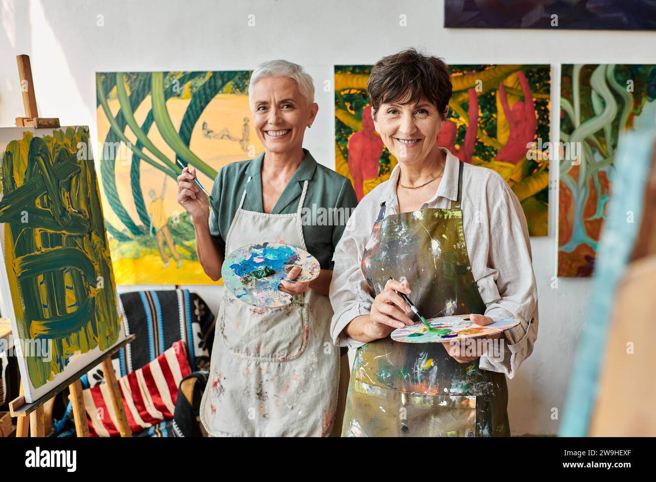 joyful mature female friends holding palettes and smiling at camera in
