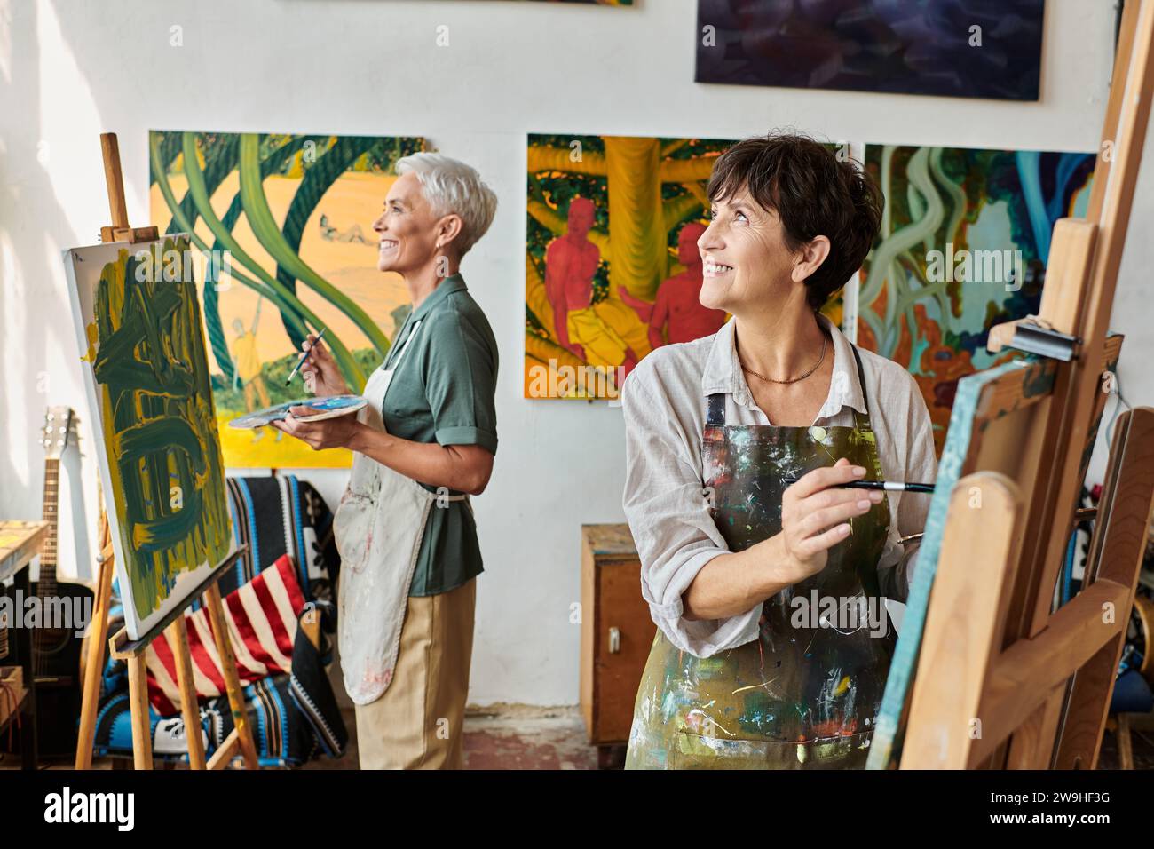 smiling mature women in aprons painting on easels and looking away in