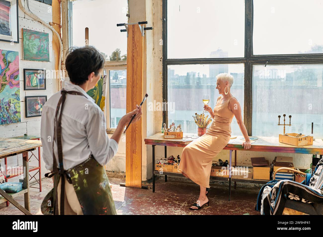 stylish mature woman posing with wine glass near female artist in light and spacious workshop Stock Photo