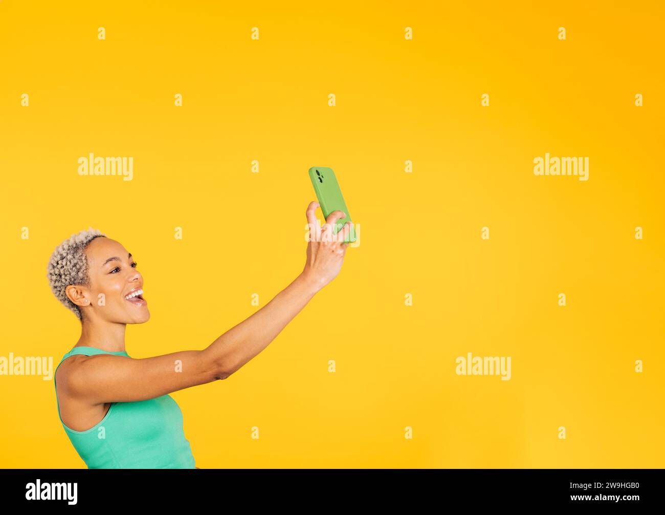 Happy young woman taking a selfie in front of a yellow background in a studio. Woman taking a selfie in a studio. Stock Photo
