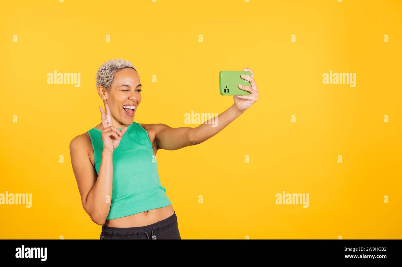 Happy young woman taking a selfie in front of a yellow background in a studio. Woman taking a selfie in a studio. Stock Photo