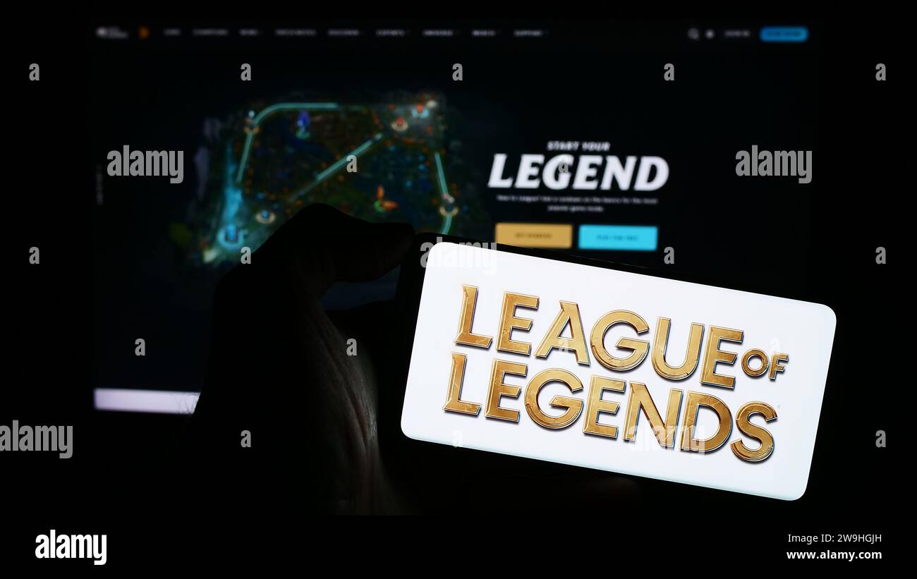 Person holding cellphone with logo of online multiplayer video game League of Legends (LoL) in front of webpage. Focus on phone display. Stock Photo