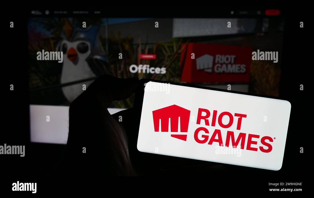 Person holding mobile phone with logo of American video game development company Riot Games Inc. in front of web page. Focus on phone display. Stock Photo