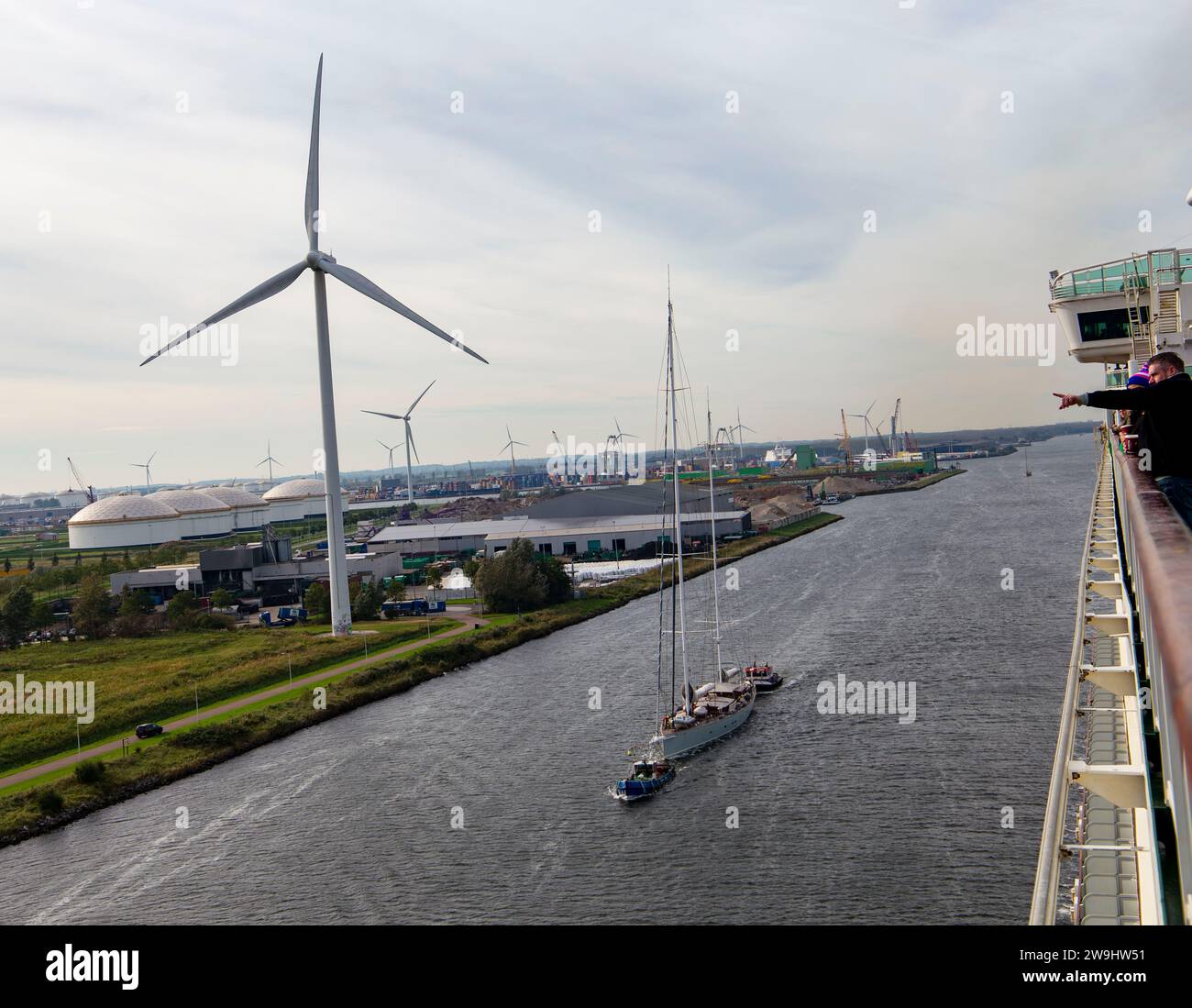 View from P&O cruise ship Ventura coming into Amsterdam with smaller sailing ship being towed by the side and electric wind turbines on shore. Stock Photo