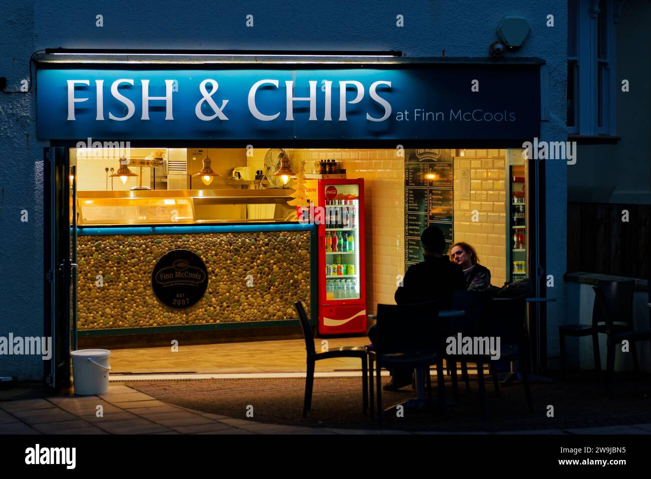 Fish and Chip shop at night with 2 people outside waiting for their order of fish and chips