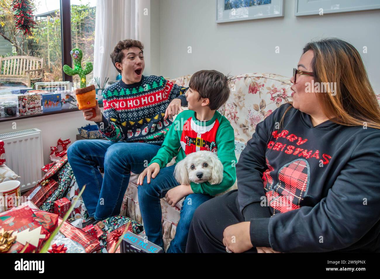 A mother and sons, sat wearing Christmas jumpers, enjoy opening Christmas presents together Stock Photo