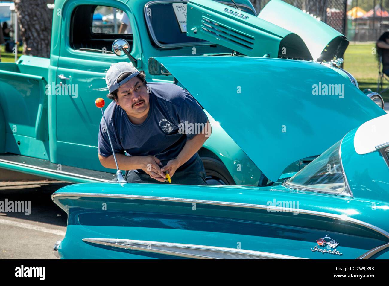 Juxtaposed with a 1940 Dodge Ram flathead truck, a Hispanic classic car collector prepares his 1958 Chevrolet Impala at a car show in Santa Ana, CA. Stock Photo