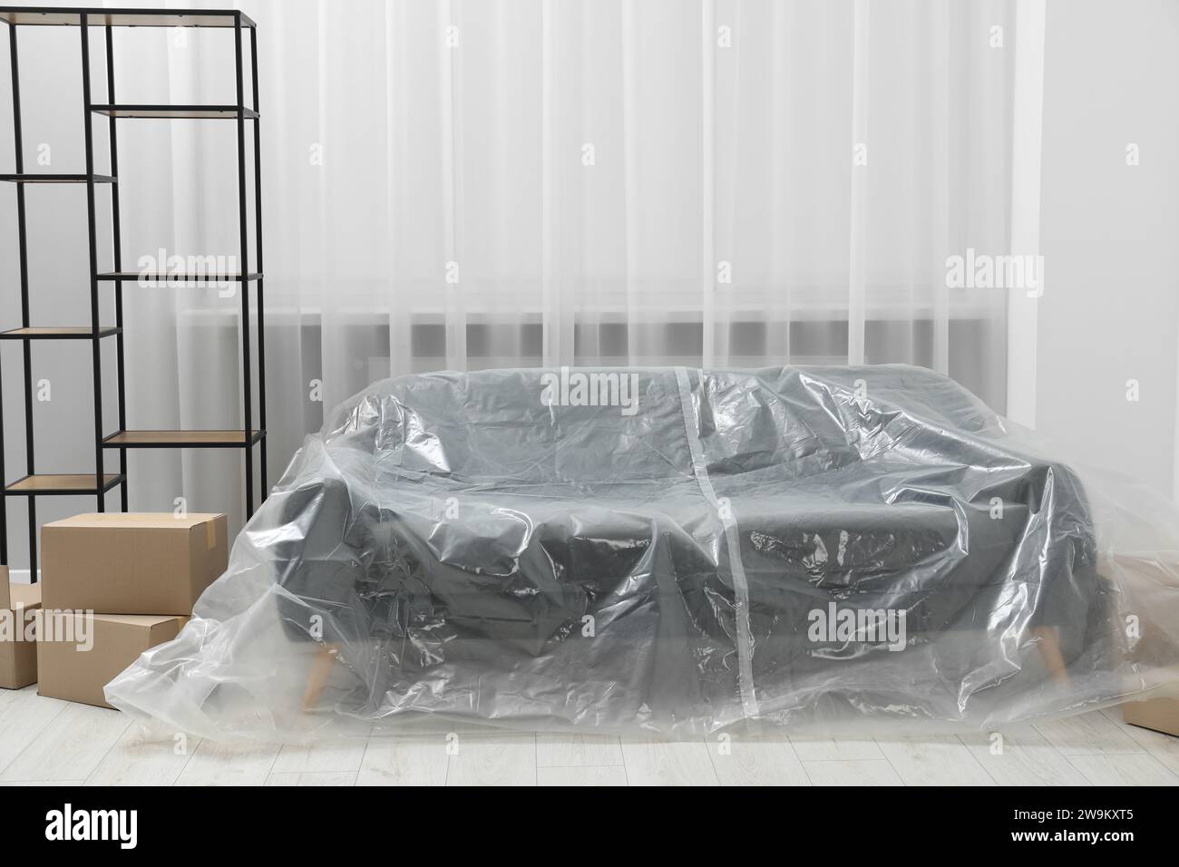 Stylish sofa covered with plastic film, shelving unit and boxes at home Stock Photo