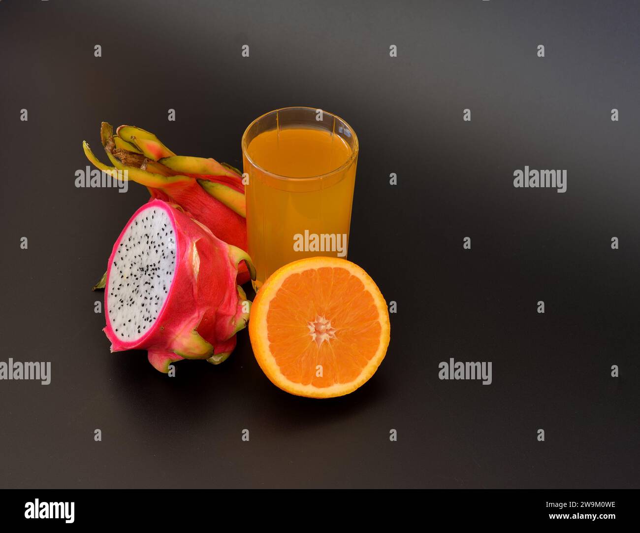 A tall glass of yellow fruit juice on a black background, next to ripe orange and pitaya fruits. Close-up. Stock Photo