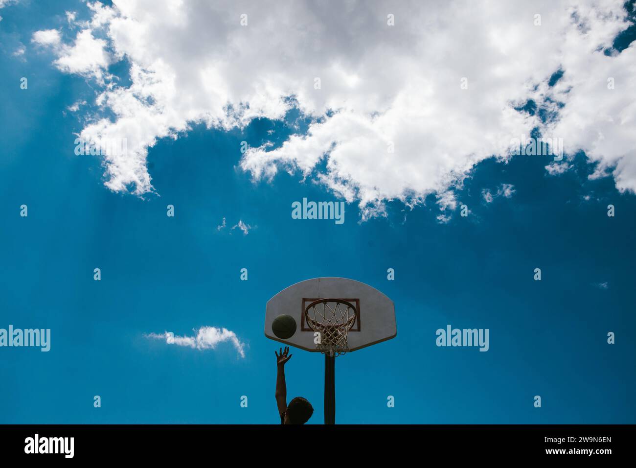 Boy making lay up at basketball hoop blue sky with clouds Stock Photo