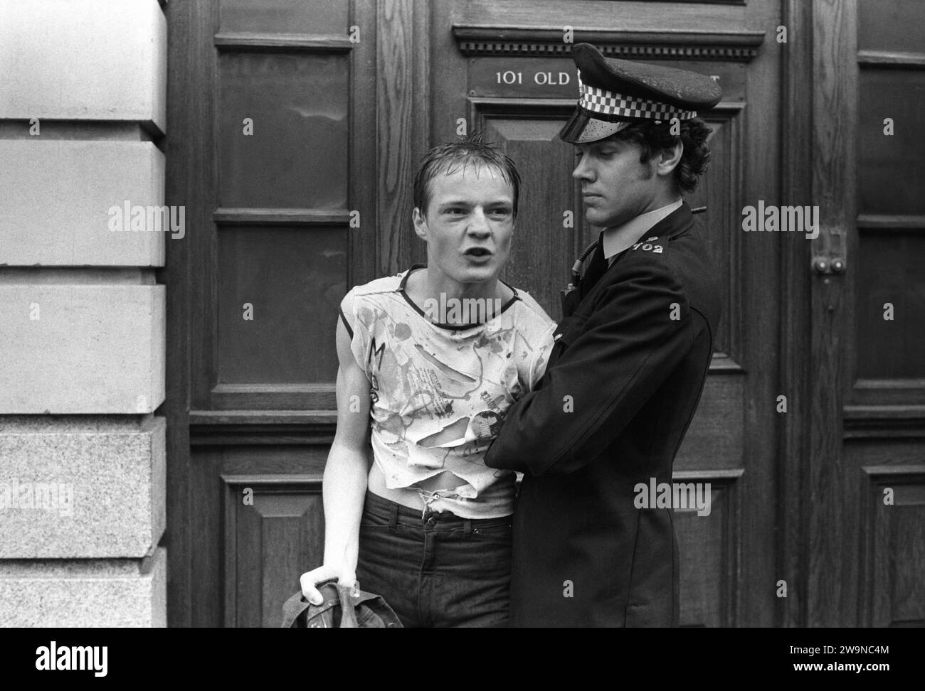 Punks UK 1970s. Punk Rocker arrest by policeman in Sloane Square Chelsea. He is wearing a fashionably torn T-shirt, neatly pinned together from Smutz, which was upstairs in Beaufort Market at 374 King’s Road. Chelsea, London, England 1977 1970s HOMER SYKES Stock Photo