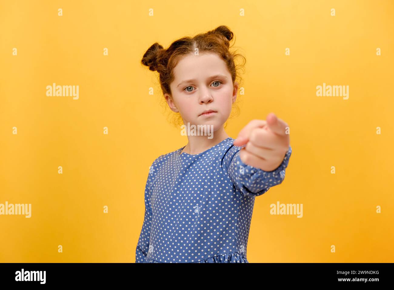 Portrait of serious little caucasian girl child pointing at camera, looking angry, making choice, choosing guilty, posing isolated over plain yellow c Stock Photo