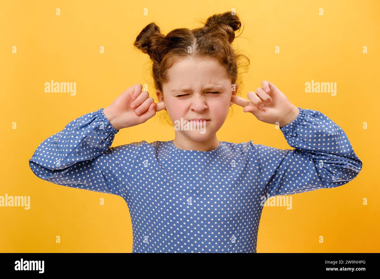 Portrait of naughty caucasian little girl child covering ears with fingers, standing isolated over plain yellow color background wall in studio with c Stock Photo