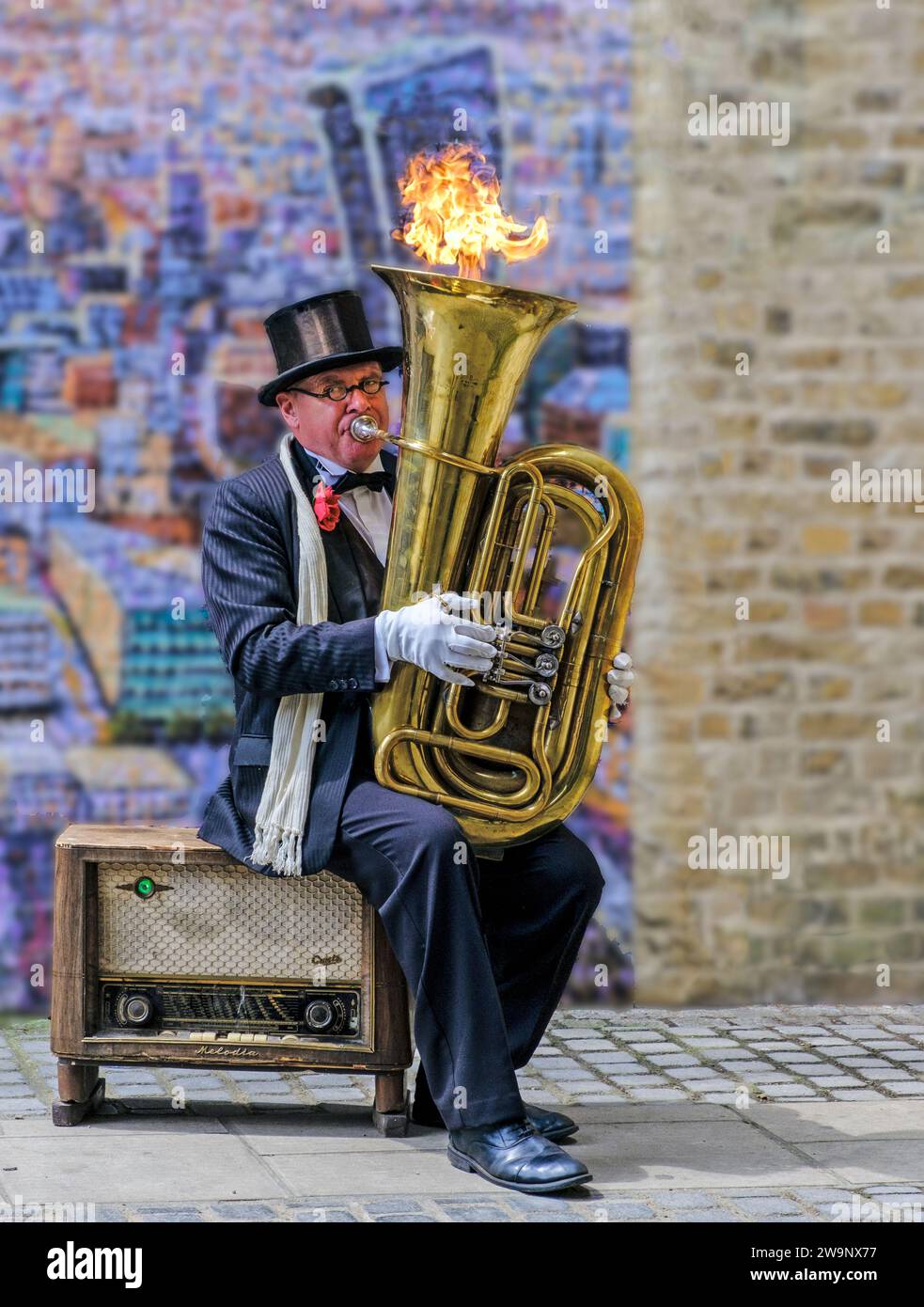 Busker Christopher Werkowicz seated on vintage radio in front of painting London Cityscape by Jimmy C, plays a tuba which shoots out fire. Stock Photo