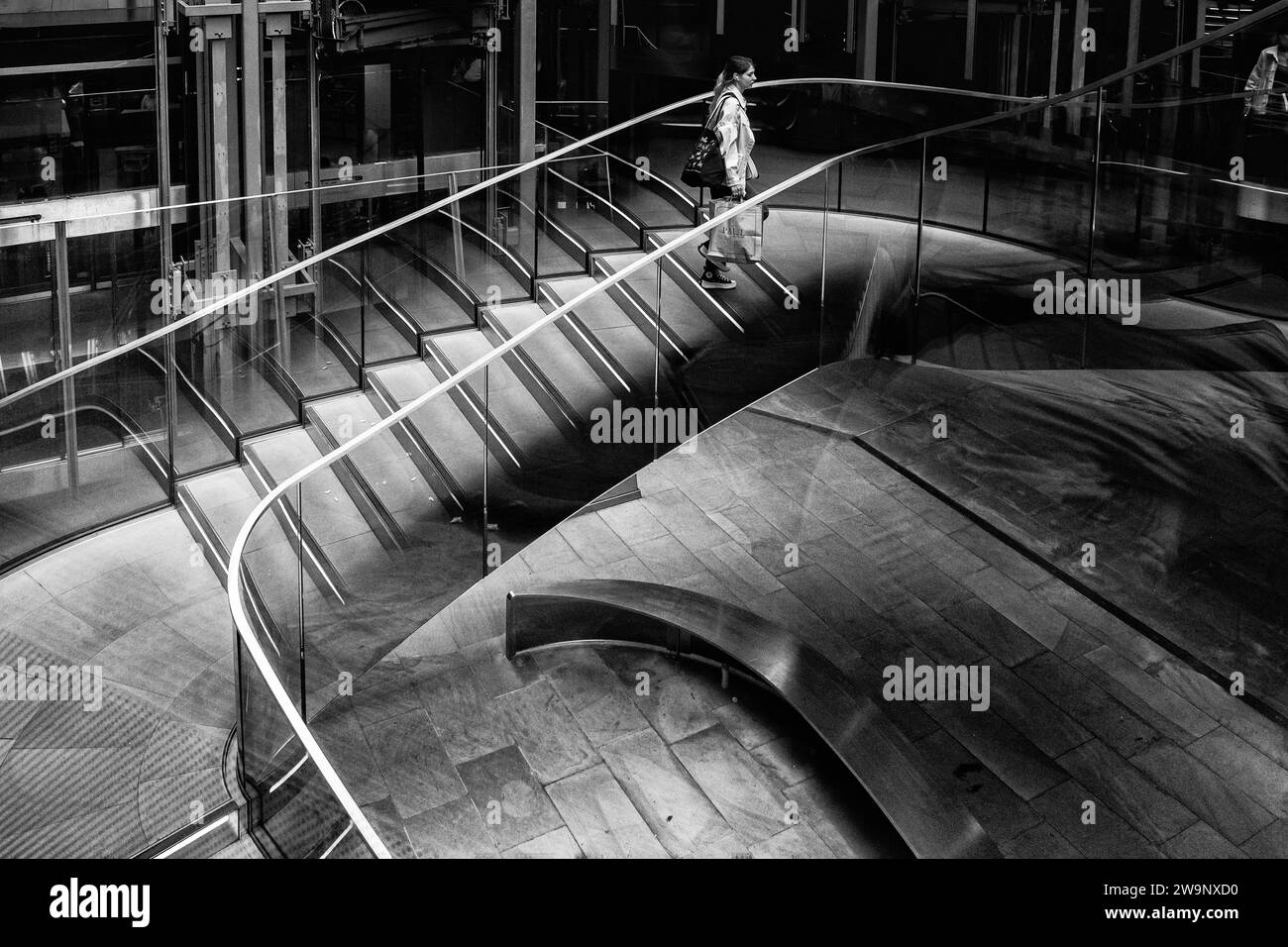 Black & white image of female holding shopping bag & walking up curved staircase Modern, indoor setting. Empty, curved metal bench on floor below. Stock Photo