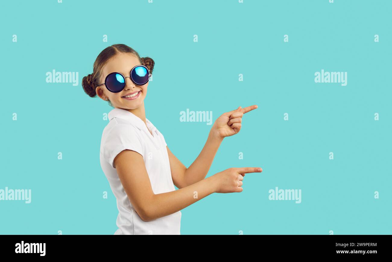 Happy child fashion model in funky sunglasses pointing to side on blue background Stock Photo
