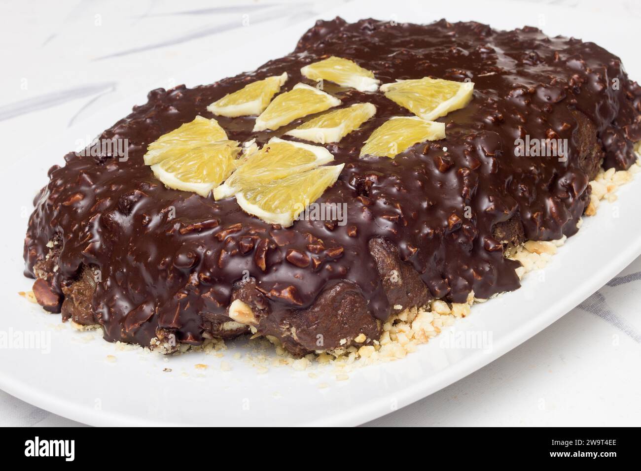 A homemade chocolate and almond delight topped with orange slices, placed on a white ceramic platter on a table covered with a tablecloth. food and ho Stock Photo