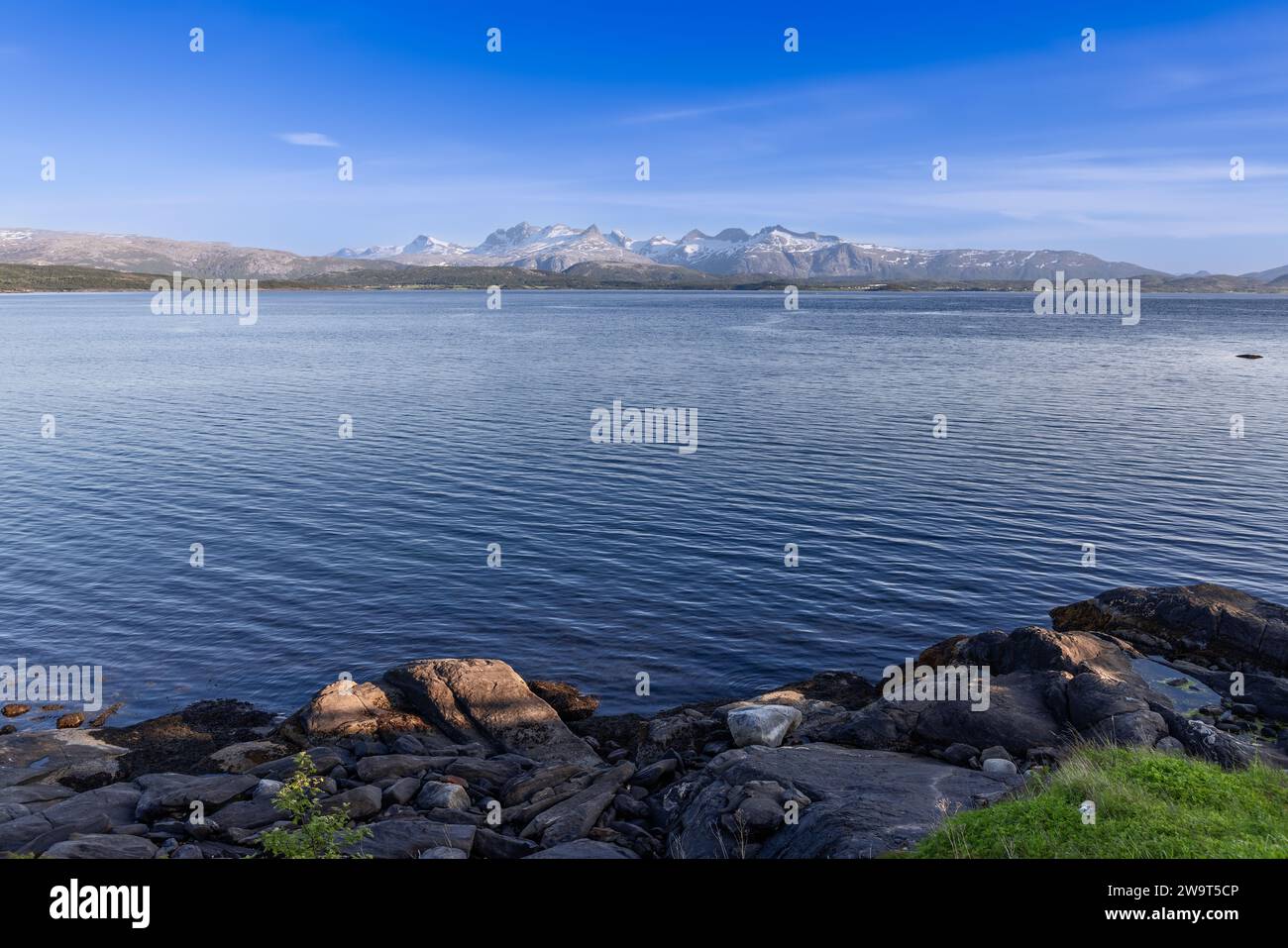 Summer view of tranquil Saltfjorden with snow-capped mountains across, large boulders in the foreground, near Bodo, Lofoten Islands, Norway Stock Photo