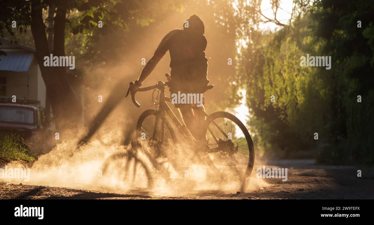 Cyclist riding a bicycle on a gravel road at sunset. A silhouette of young sporty man on a gravel bike in a cloud of dust from real whell after skiddi Stock Photo