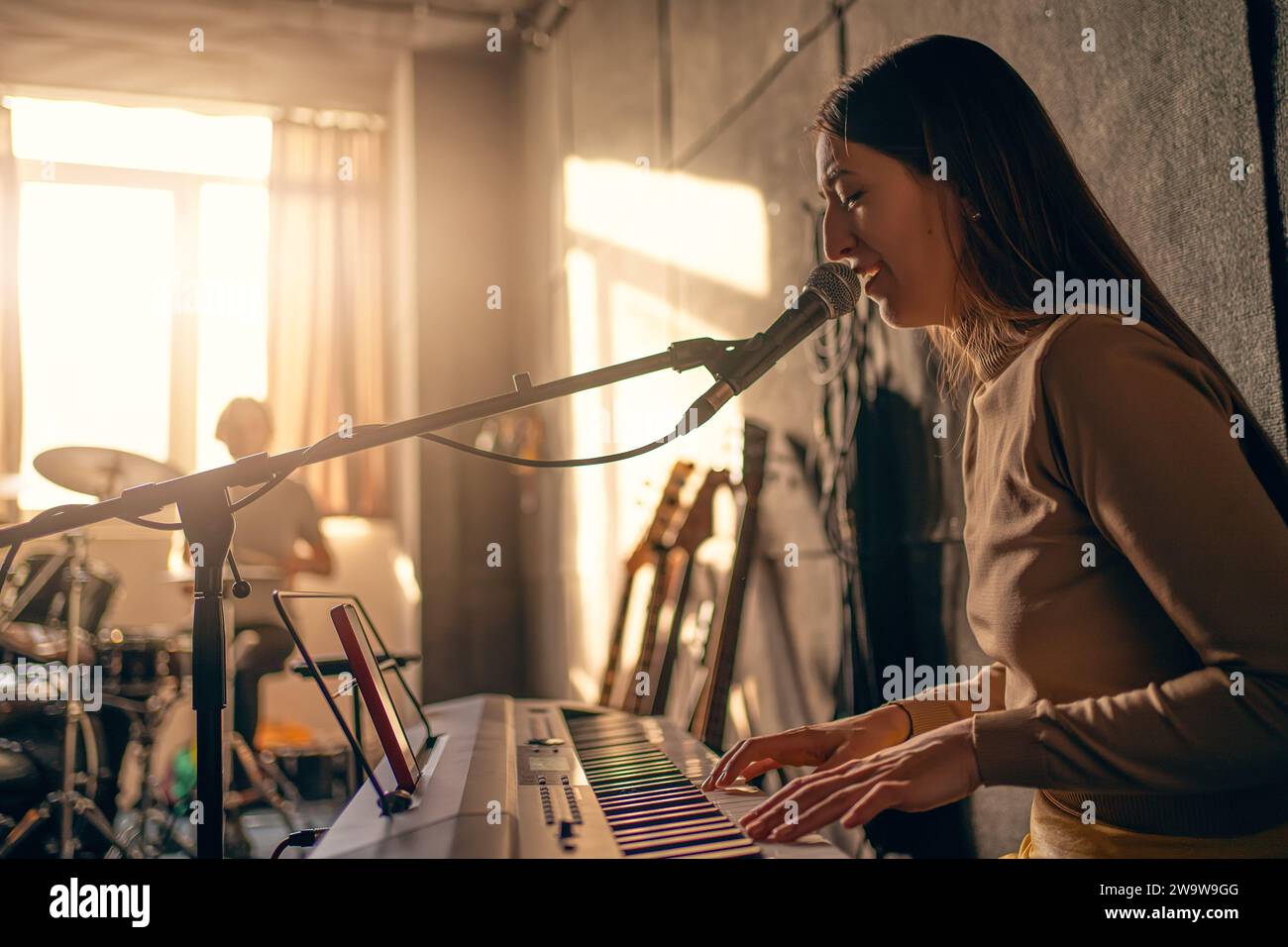 Girl singer plays synthesizer and sings into microphone in music studio. Music band rehearsal. Stock Photo