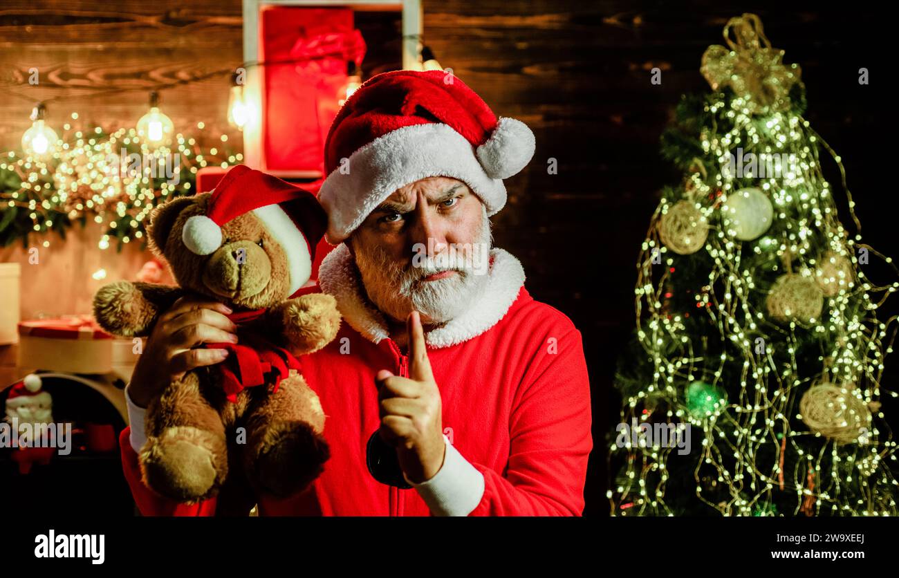 Serious Santa Claus with plush teddy bear in room decorated for Christmas. New Year holidays. Christmas background. Bearded man in Santa costume with Stock Photo