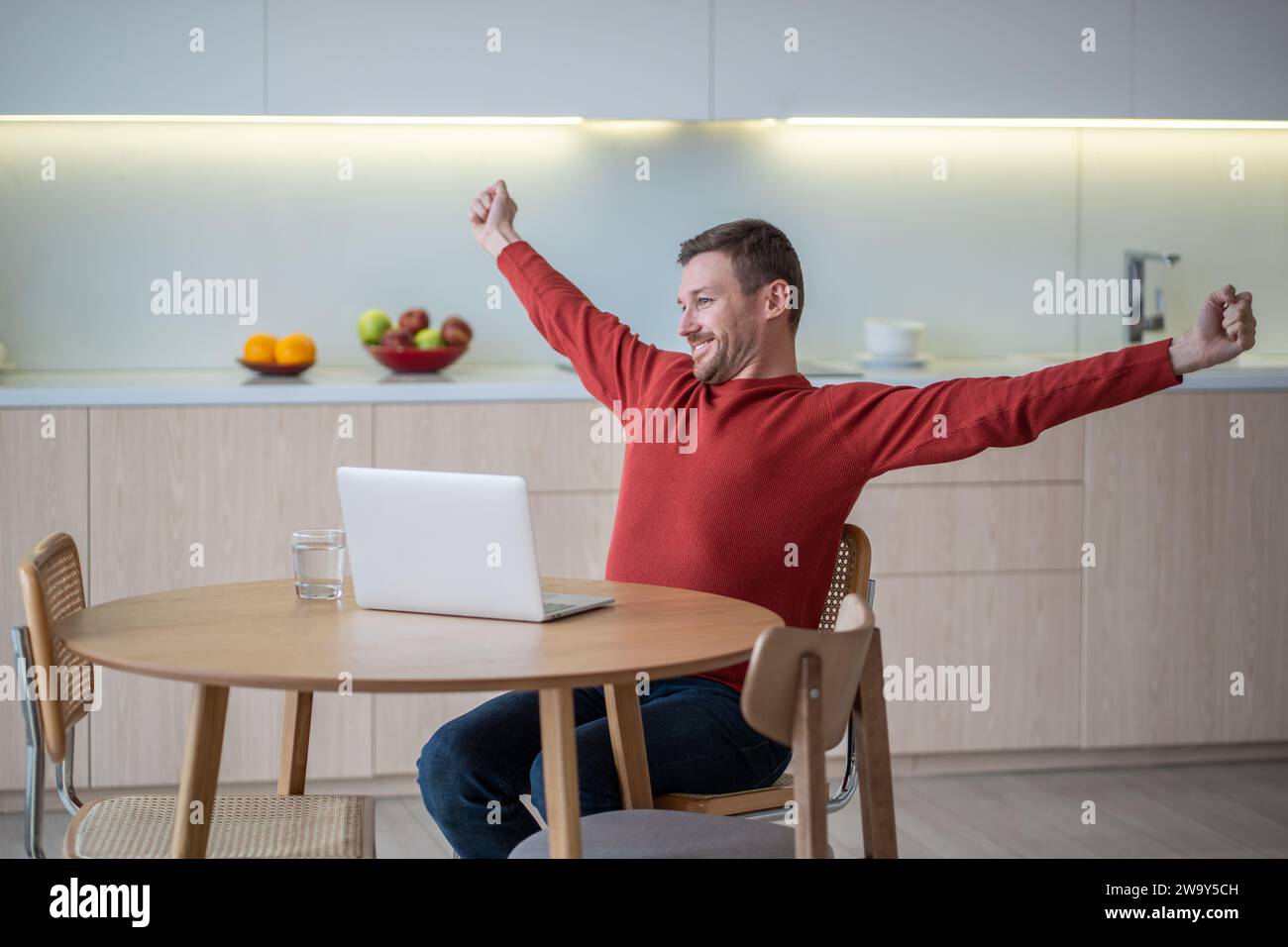 Happy freelancer man stretching arms smiling ending work sitting on kitchen with laptop.  Stock Photo