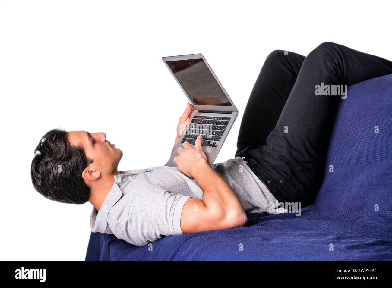 Young attractive man working with computer while laying in awkward position on couch, on white background Stock Photo