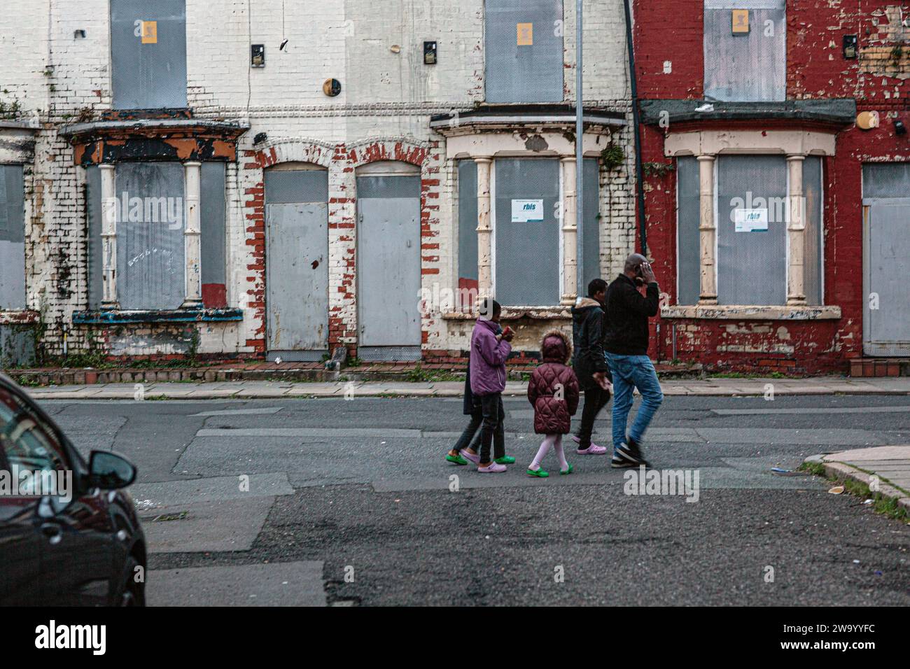 Family of immigrants passing rundown boarded Up, sealed and empty Terraced Property, house, terrace in Liverpool . Stock Photo