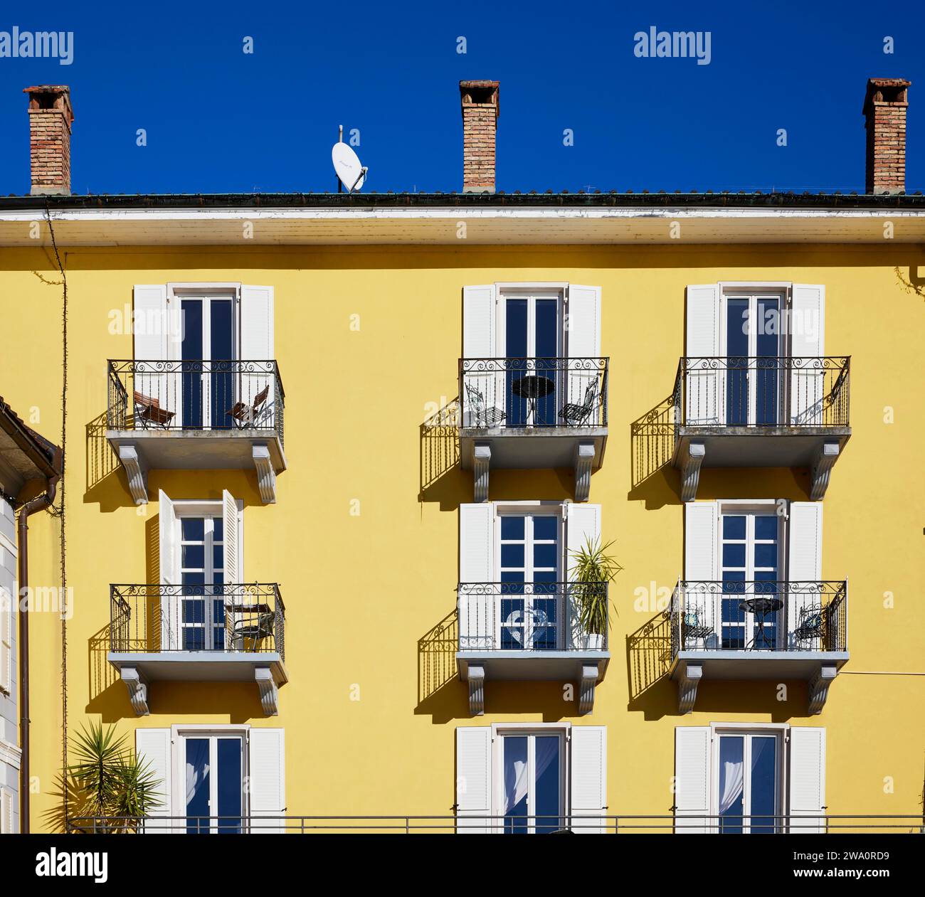 Light yellow façade with windows, white shutters, balconies with wrought iron grilles and palm trees against a deep blue sky in Ascona, district of Lo Stock Photo